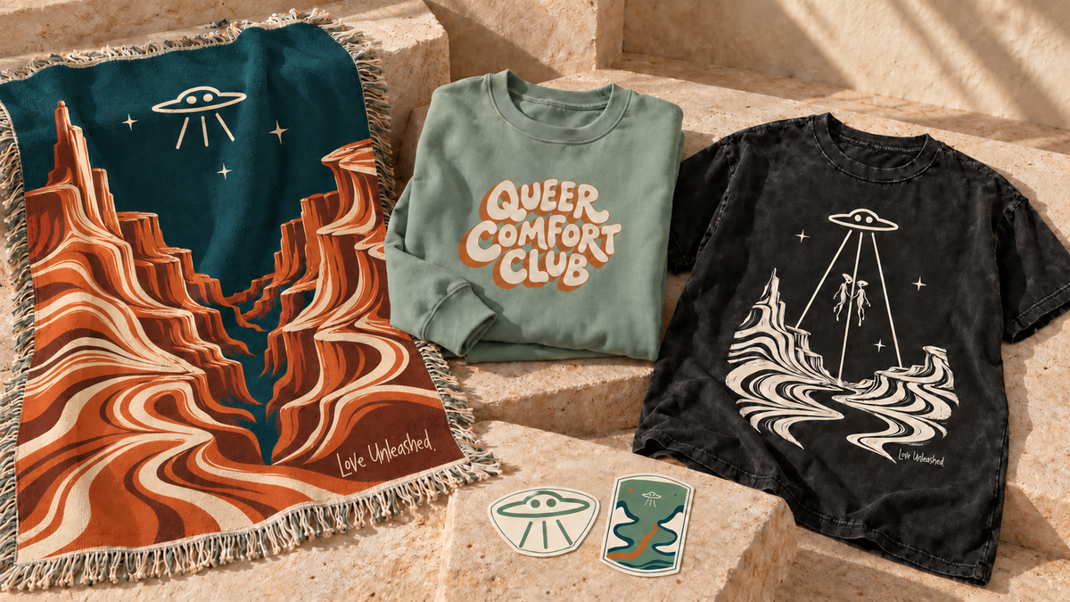 A Love Unleashed collection scene with a night-canyon throw, Queer Comfort Club crewneck, alien-pair tee, and two stickers